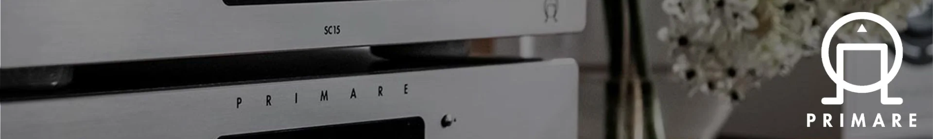 A sleek silver audio component with "PRIMARE" branding and the model "SC15" is stacked in a modern setting. Flowers are softly blurred in the background.