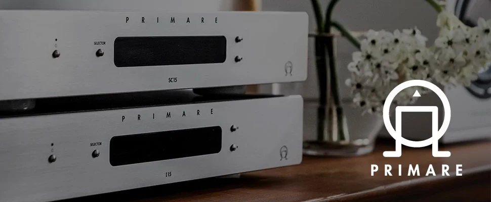 A sleek silver audio component with "PRIMARE" branding and the model "SC15" is stacked in a modern setting. Flowers are softly blurred in the background.