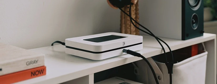 A sleek, white audio streaming device sits on a white shelf, connected by black cables. Nearby are books and a speaker, conveying a modern, tech-savvy vibe.