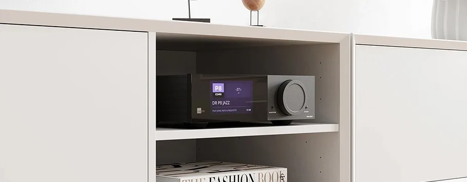 A sleek black stereo receiver with a digital display sits on a white shelf. A book titled "The Fashion Book" is below it, creating a modern, minimalist vibe.
