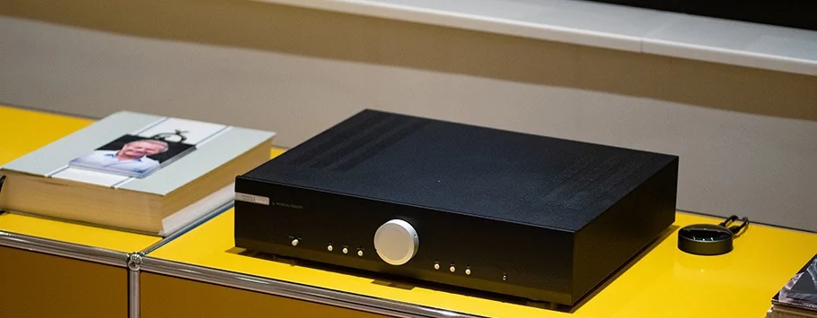 A black audio amplifier with a large silver dial sits on a yellow shelf next to a closed book with a bookmark, creating a sleek, modern aesthetic.
