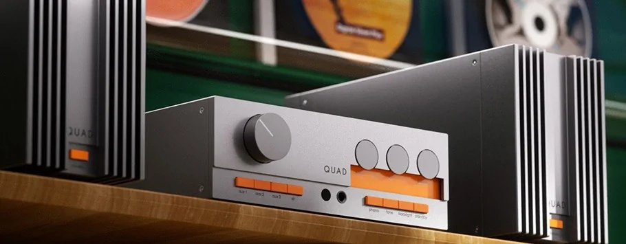 Silver audio equipment with orange LED displays sits on a wooden shelf. The setup includes a large knob and multiple buttons. The background shows framed records.