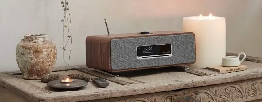 Vintage wooden radio with a digital display sits on a rustic carved table. Surrounding it are a weathered vase, lit candles, and a cup, creating a cosy ambiance.