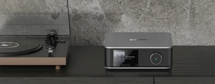 A sleek turntable and modern music player displayed on a dark wooden surface against a textured grey wall. The music player screen shows an album cover.