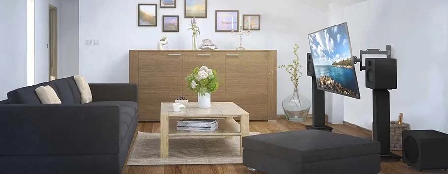 A modern living room with a dark grey sofa, beige cushions, and a light wood coffee table. A large television and speakers are set up opposite. The wooden cabinet in the background holds framed pictures, vases, and candles, creating an inviting atmosphere