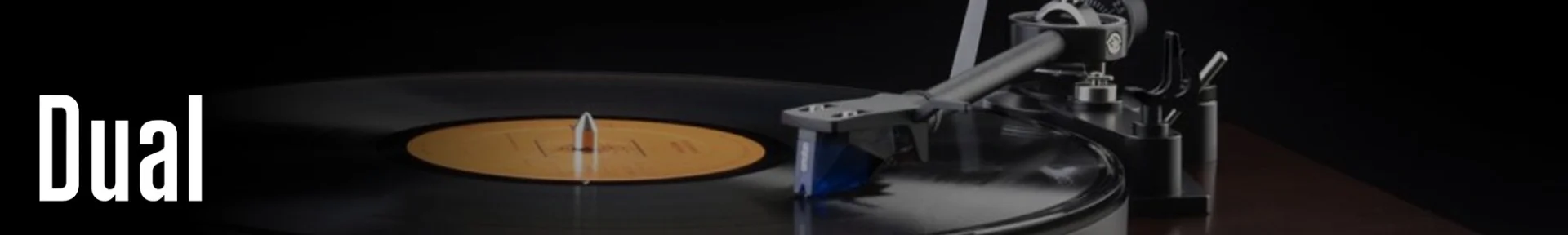 Turntable playing a vinyl record with a yellow label, set on a dark wooden surface. The word "Dual" appears in white on the left side.