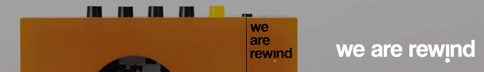 Close-up of an orange retro-style cassette player with black and yellow buttons on top. The text "we are rewind" is printed in black on the device. The background is grey.