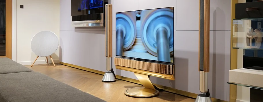 Modern living room with a sleek TV on a gold stand, flanked by stylish speakers. A round speaker sits in the corner, creating a sophisticated tech vibe.