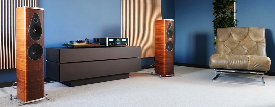 A modern audio setup in a room with blue walls and acoustic panels. Two tall, wooden speakers flank a sleek black console. A soft leather chair adds comfort.