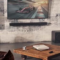 A modern living room setup featuring a large flat-screen TV displaying a Formula 1 race above a wall-mounted soundbar, with a rustic wooden coffee table in the foreground holding books and a small cup.