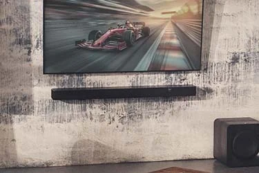 A modern living room setup featuring a large flat-screen TV displaying a Formula 1 race above a wall-mounted soundbar, with a rustic wooden coffee table in the foreground holding books and a small cup.