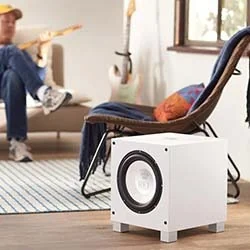 A modern white subwoofer sits on a wooden floor, while a man in casual attire plays guitar in a cosy living room setting.