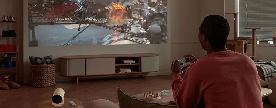 Person playing a video game on a large projection screen in a cosy living room. The room features warm lighting, wooden furniture, and a chessboard.