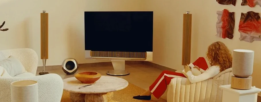 A cozy living room features a modern TV and tall speakers. A person with curly hair and red-striped pants sits in a cream chair, creating a relaxed atmosphere.