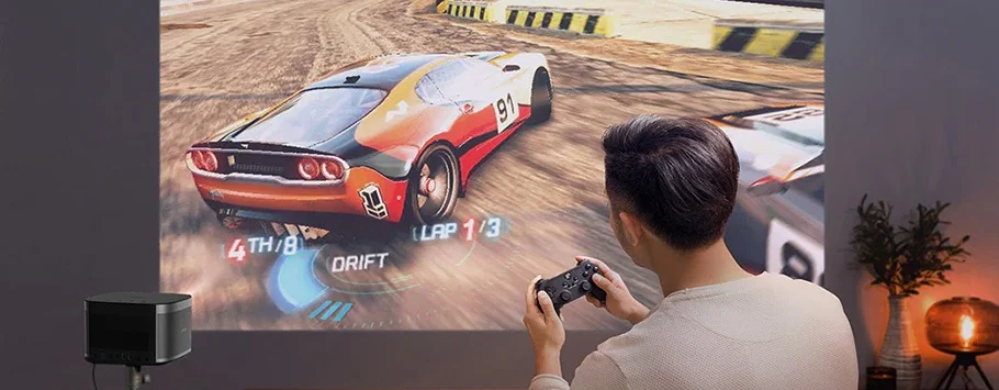 A person plays a racing video game projected on a wall. The game shows a car drifting on a track. The setup includes a small projector and cosy lighting.