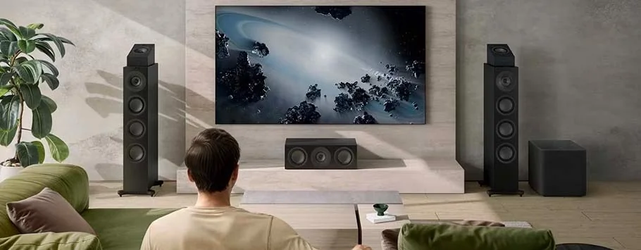 A person sits on a couch in a modern living room, watching a large TV showing a space scene. Tall speakers flank the TV, creating a cinematic atmosphere.