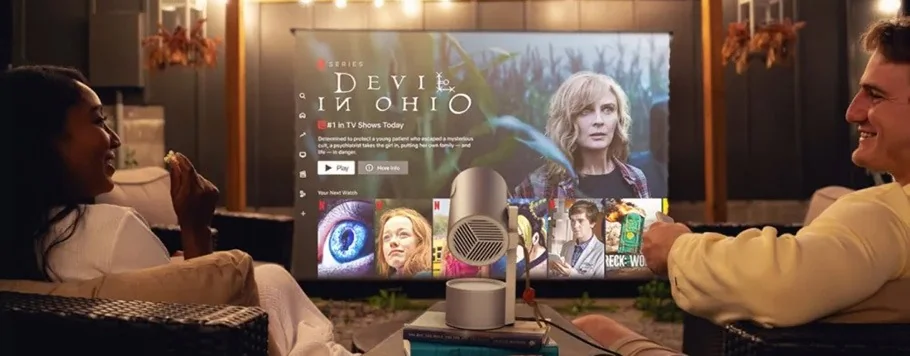 A man and woman smile while watching a Netflix show, "Devil in Ohio," outdoors at night. A mini projector displays the TV screen. Cosy atmosphere.
