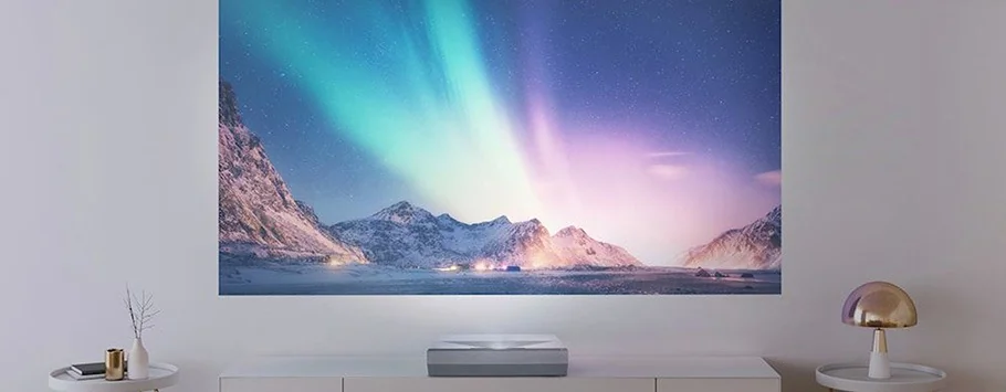 A projector casts a vivid aurora borealis scene on a wall, showcasing colourful lights above snowy mountains. Minimalist décor includes tables with a lamp and vase.