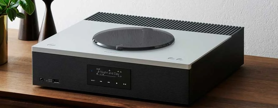 A sleek black audio CD player sits atop a wooden cabinet with a modern, minimalist design. Soft lighting casts gentle shadows.