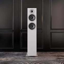 Tall white speaker with three circular drivers stands on a patterned rug against a dark-paneled wall. The setting is modern and sleek.