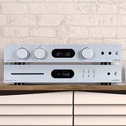 Silver stereo system with digital displays and control knobs, placed on a wooden shelf against a textured white brick wall. Sleek and modern design.