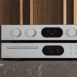 Silver stereo amplifier and CD player with digital displays and round control knobs, set against a wooden-panel background. Modern and sleek design.