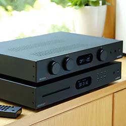 Stacked black stereo components on a wooden table with control knobs and digital displays, accompanied by a remote. Bright, modern setting with soft light.