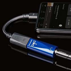 A blue audio adapter connected to a smartphone on a reflective black surface. The phone displays a music player app, suggesting a high-quality audio setup.