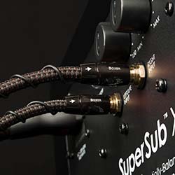 Close-up of audio cables plugged into a black audio device labeled "SuperSub X." The braided cables and sleek knobs suggest a high-quality, engineered design.