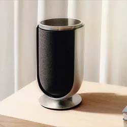 A sleek, modern speaker with a black fabric front and metallic sides stands on a wooden table. Soft natural light filters through a curtain behind it.