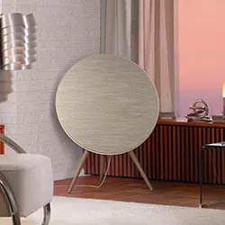 A stylish modern room with a circular speaker on tripod legs against a white brick wall. Soft pink sunset visible through a window adds warmth.