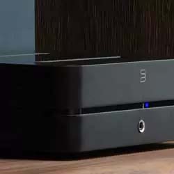 Close-up of a sleek black audio amplifier on a wooden surface. The device features a minimalistic design with a subtle blue indicator light.