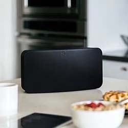 A sleek black speaker sits on a kitchen counter, surrounded by a mug, phone, and bowl of cereal. The setting is modern and tidy, conveying a relaxed ambiance.