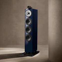 A sleek, tall blue speaker with four circular drivers stands in a minimalistic room. The setup exudes modernity and elegance, casting a soft shadow.