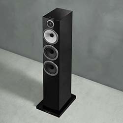A sleek black floor-standing speaker with three silver circular drivers is positioned on a light gray floor. The background is a textured gray wall.