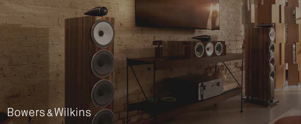 Living room with two Bowers & Wilkins speakers and a brown media console on a brick wall. Warm lighting creates a cozy, sophisticated atmosphere.