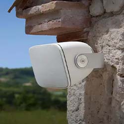 A modern white security camera is mounted on a stone wall under a wooden beam, overlooking a lush, green landscape and blue sky. The scene feels serene and secure.
