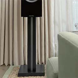 A black speaker on a tall stand is in a cozy living room with cream curtains and a light green sofa, creating a calm and elegant atmosphere.