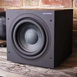 A black subwoofer sits on a wooden surface against a brick wall. The speaker cone is prominently visible, emphasizing a modern, minimalist design.