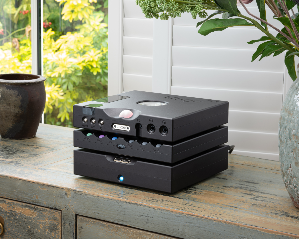 Stacked black audio equipment sits on a vintage wooden table. Nearby are a ceramic pot and potted plant. Sunlight streams in through white shutters.