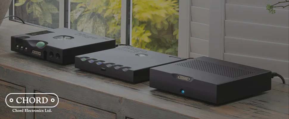 Three sleek audio devices from Chord Electronics are displayed on a wooden surface near a window with greenery outside, conveying a modern and sophisticated tone.