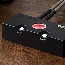 A black audio input device on a wooden table features buttons labeled "Filter" and "Input." Cables extend from the back, and a red light is visible.