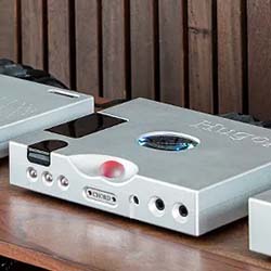 Three sleek, silver audio components with knobs and ports are placed on a wooden shelf. The design is modern, with a minimalist aesthetic.