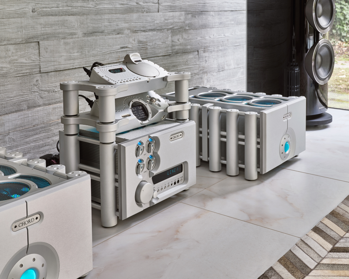 High-end audio equipment setup featuring sleek silver components with glowing blue accents, set on a polished floor against a textured gray wall.