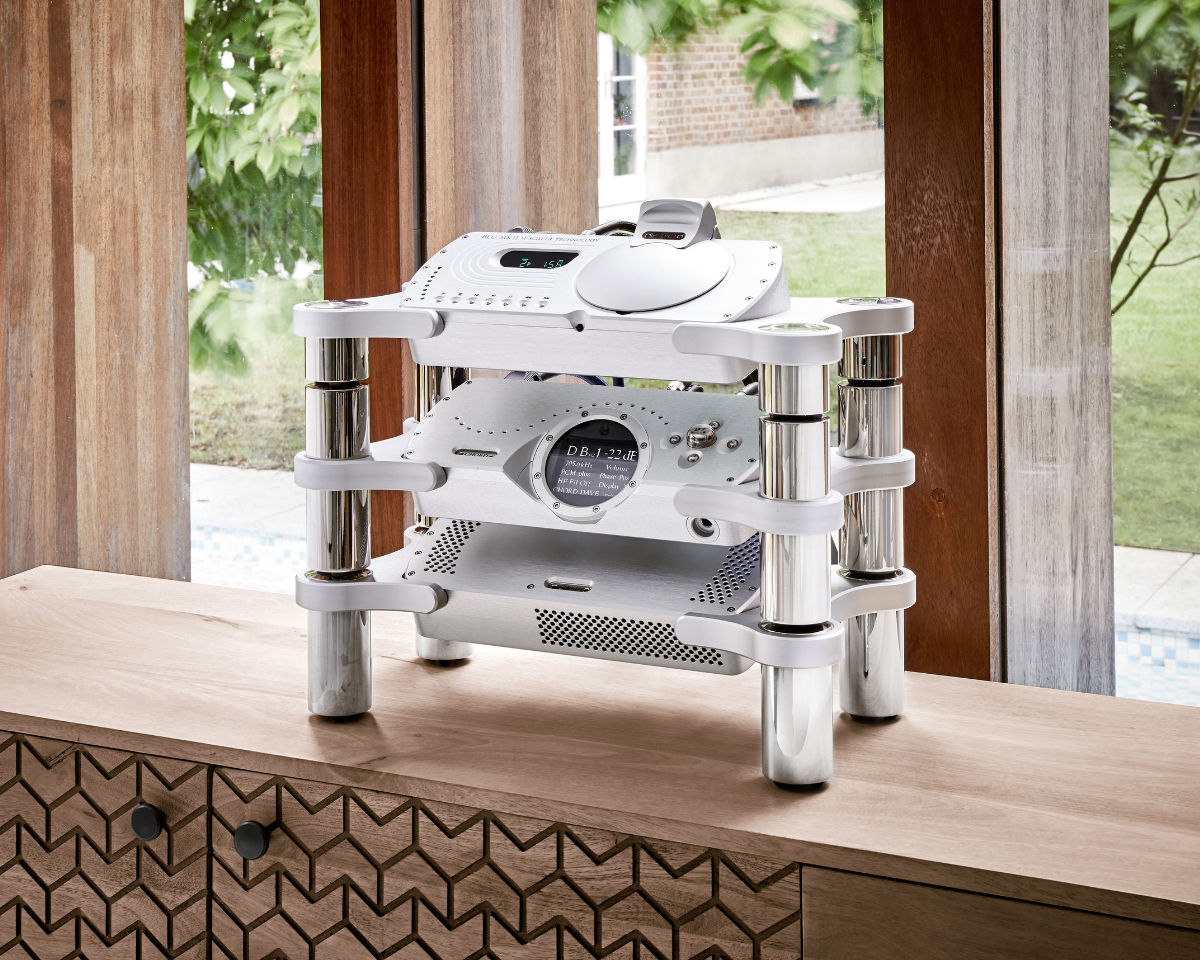 A sleek, modern audio system with a white and metallic design sits on a wooden cabinet. Behind it, large windows reveal a lush, green garden.