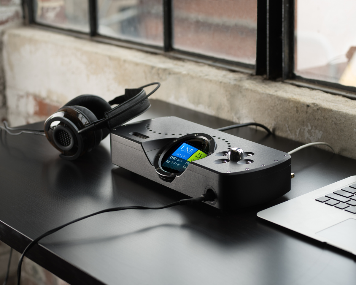Headphones rest beside a sleek audio device with a digital display showing USB input. They're on a minimalist black desk near a window.