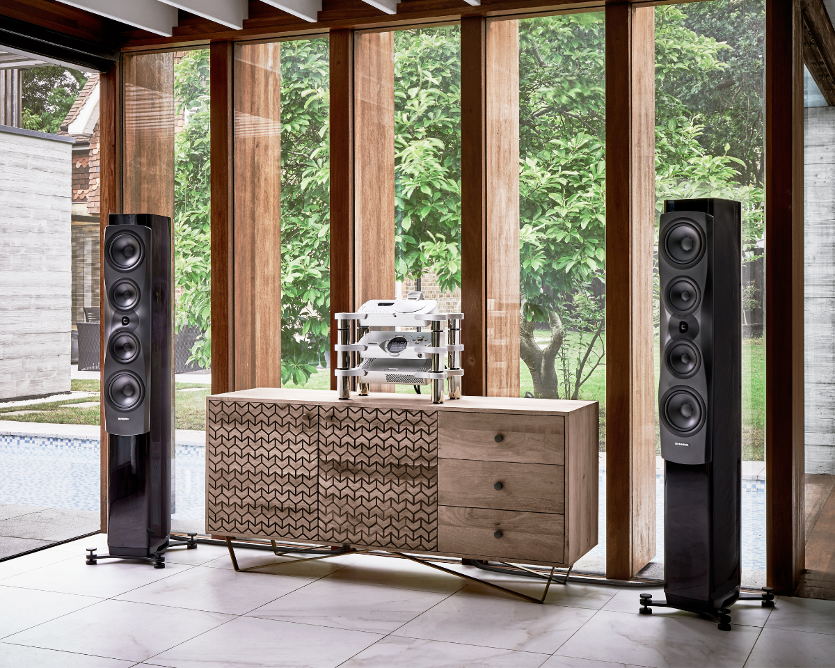 Modern living room with two tall, sleek black speakers flanking a wooden cabinet with intricate patterns. Large windows reveal lush green trees outside.
