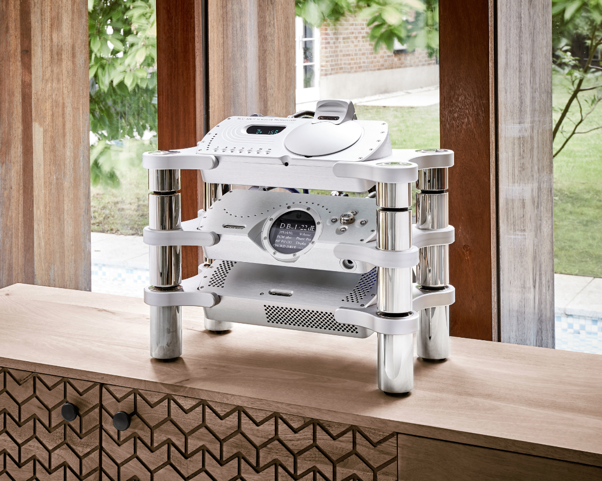 A sleek silver audio equipment stack with metal supports sits on a wooden cabinet. Sunlight filters through large windows, revealing greenery outside.