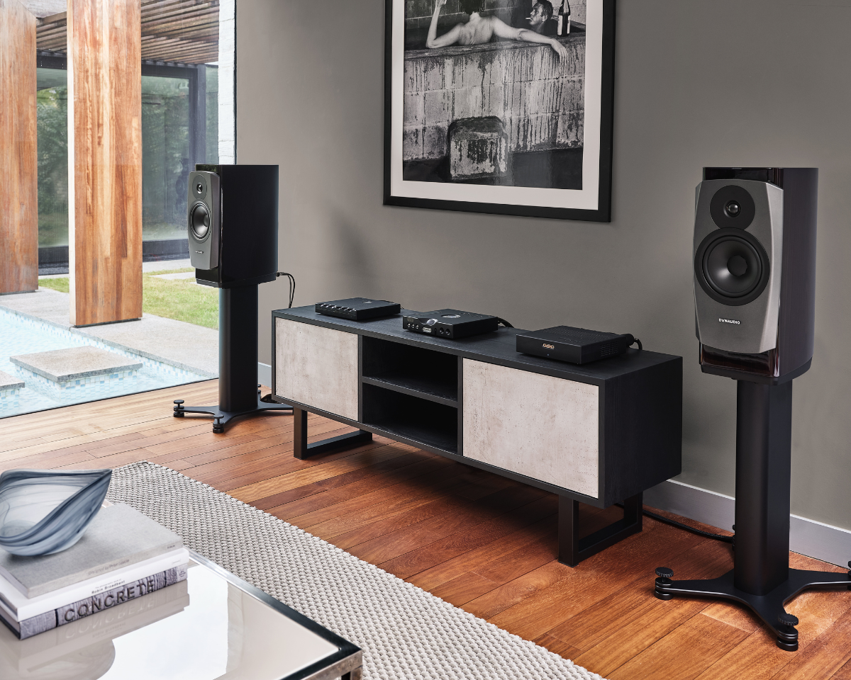 A modern living room with large speakers on stands flanking a sleek media console. A black-and-white photo hangs above, and a pool is visible outside.