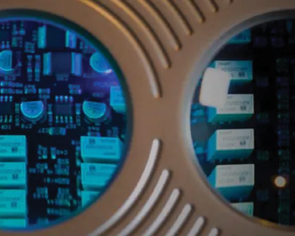 A close-up view of a circuit board through two circular windows on a metallic surface. Blue and teal lights reflect off the electronic components, creating a futuristic feel.